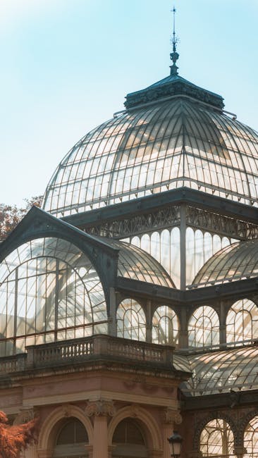 Photograph of a glass conservatory with tall, arched windows supported by ornate, cream-coloured frames and columns, featuring decorative painted details along the lower walls. Inside the conservatory