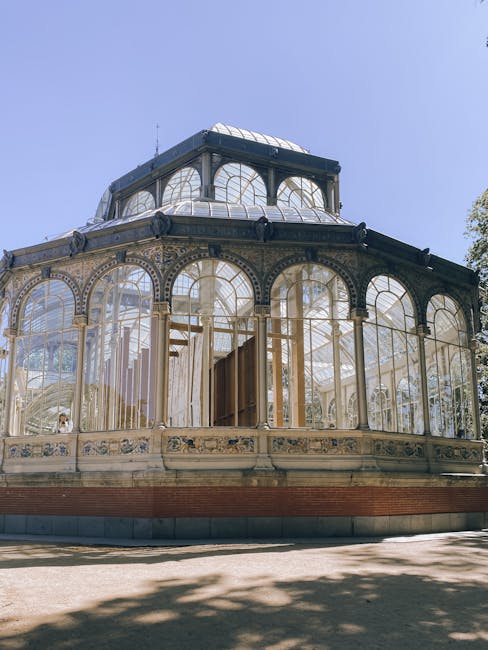 A historic glass conservatory with ornate metal framing and large arched windows, located outdoors on a sunny day with clear blue sky above. The structure is surrounded by trees casting shadows on the ground, and its interior is visible through the glass panes, showing wooden doors and some construction or maintenance activity. The conservatory is part of a property space used for house removals and furniture transport, with the setting suggesting a scheduled move or relocation process managed by Man and a Van Crystal Palace. The scene highlights the importance of careful handling of fragile items such as large glass and metal objects during home relocation and packing and moving services in the Crystal Palace area.