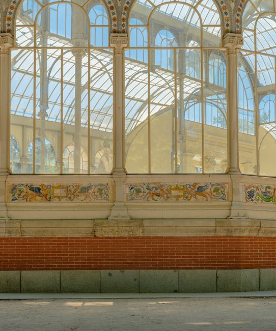 A historic glass conservatory with ornate metal framing and large arched windows, located outdoors on a sunny day with clear blue sky above. The structure is surrounded by trees casting shadows on the ground, and its interior is visible through the glass panes, showing wooden doors and some construction or maintenance activity. The conservatory is part of a property space used for house removals and furniture transport, with the setting suggesting a scheduled move or relocation process managed by Man and a Van Crystal Palace. The scene highlights the importance of careful handling of fragile items such as large glass and metal objects during home relocation and packing and moving services in the Crystal Palace area.