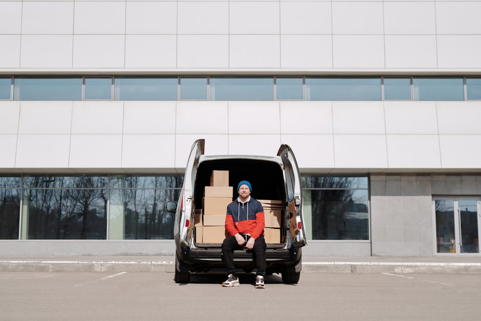 A man wearing a blue beanie, red and navy jacket, and black pants is seated on the open tailgate of a black van positioned outside a modern building with large glass windows and a grey facade. The van is loaded with several cardboard boxes of various sizes, some stacked on top of each other, indicating the process of furniture transport during a home relocation or packing and moving operation. The boxes are secured with packing materials such as plastic wrap or cardboard. The van is parked on a paved area, possibly a designated unloading zone, with no other vehicles visible nearby. The surrounding environment includes leafless trees reflected in the building's windows, suggesting a winter season. This scene exemplifies the loading process involved in professional removals services, supported by [COMPANY_NAME], focusing on efficient house moves and furniture transport, with ample space for ease of unloading and planning parking for smooth relocation logistics.