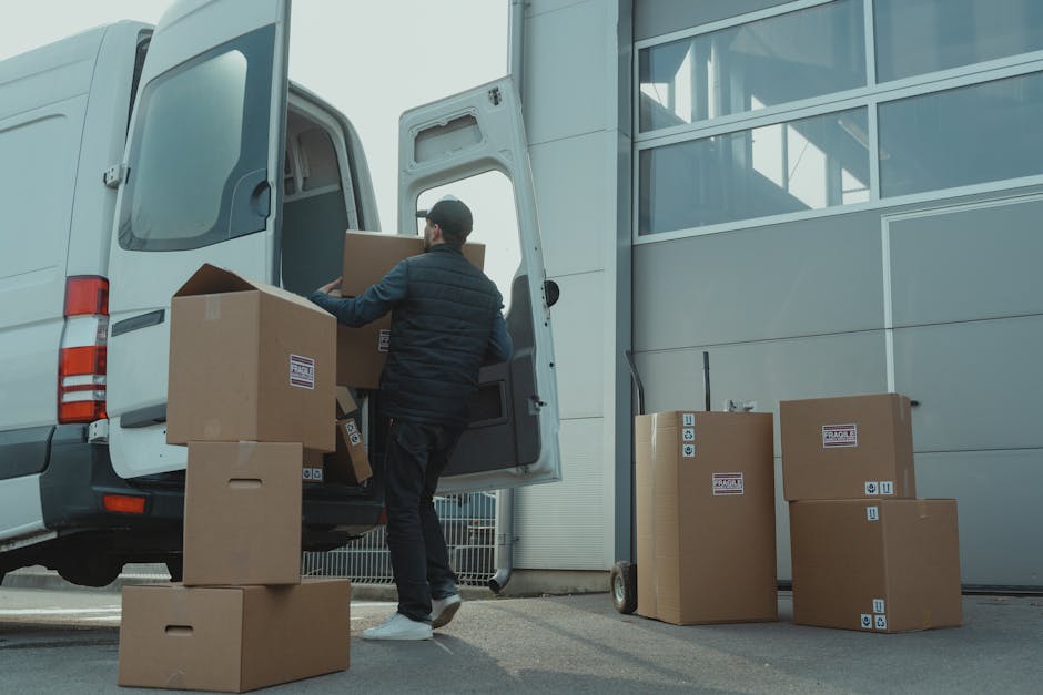 A man dressed in dark clothing, including a black beanie and a navy blue jacket, is loading large brown cardboard boxes into the back of a white moving van parked outside a modern building with large glass windows. The boxes are packed with lightweight packing materials, some wrapped in plastic, and are labeled with handling symbols and fragile stickers. Several boxes, some on a small trolley, are standing upright on the pavement nearby, ready for transport. The loading process involves the use of a trolley for ease of movement, with the man carrying boxes towards the open rear door of the van, which features an internal floor and side partitions to secure items during transit. The environment is well-lit, and the scene depicts an organized home relocation or furniture transport operation coordinated by Man and a Van Crystal Palace as part of their removals service, emphasizing efficient packing and loading practices suited for house removals in the CR19 area.