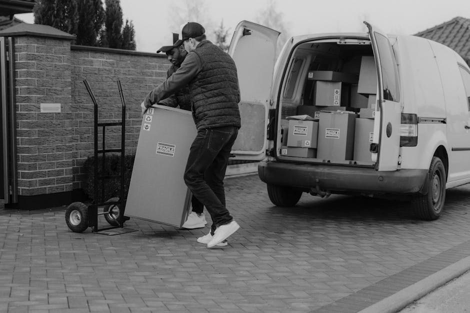 A man wearing a blue beanie, red and navy jacket, and black pants is seated on the open tailgate of a black van positioned outside a modern building with large glass windows and a grey facade. The van is loaded with several cardboard boxes of various sizes, some stacked on top of each other, indicating the process of furniture transport during a home relocation or packing and moving operation. The boxes are secured with packing materials such as plastic wrap or cardboard. The van is parked on a paved area, possibly a designated unloading zone, with no other vehicles visible nearby. The surrounding environment includes leafless trees reflected in the building's windows, suggesting a winter season. This scene exemplifies the loading process involved in professional removals services, supported by [COMPANY_NAME], focusing on efficient house moves and furniture transport, with ample space for ease of unloading and planning parking for smooth relocation logistics.