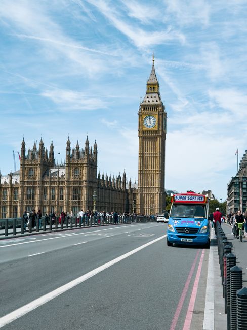 The image shows a street scene near the Palace of Westminster in London, with the iconic Elizabeth Tower and Big Ben clock visible in the background. On the road, there is a blue Mercedes-Benz van wit